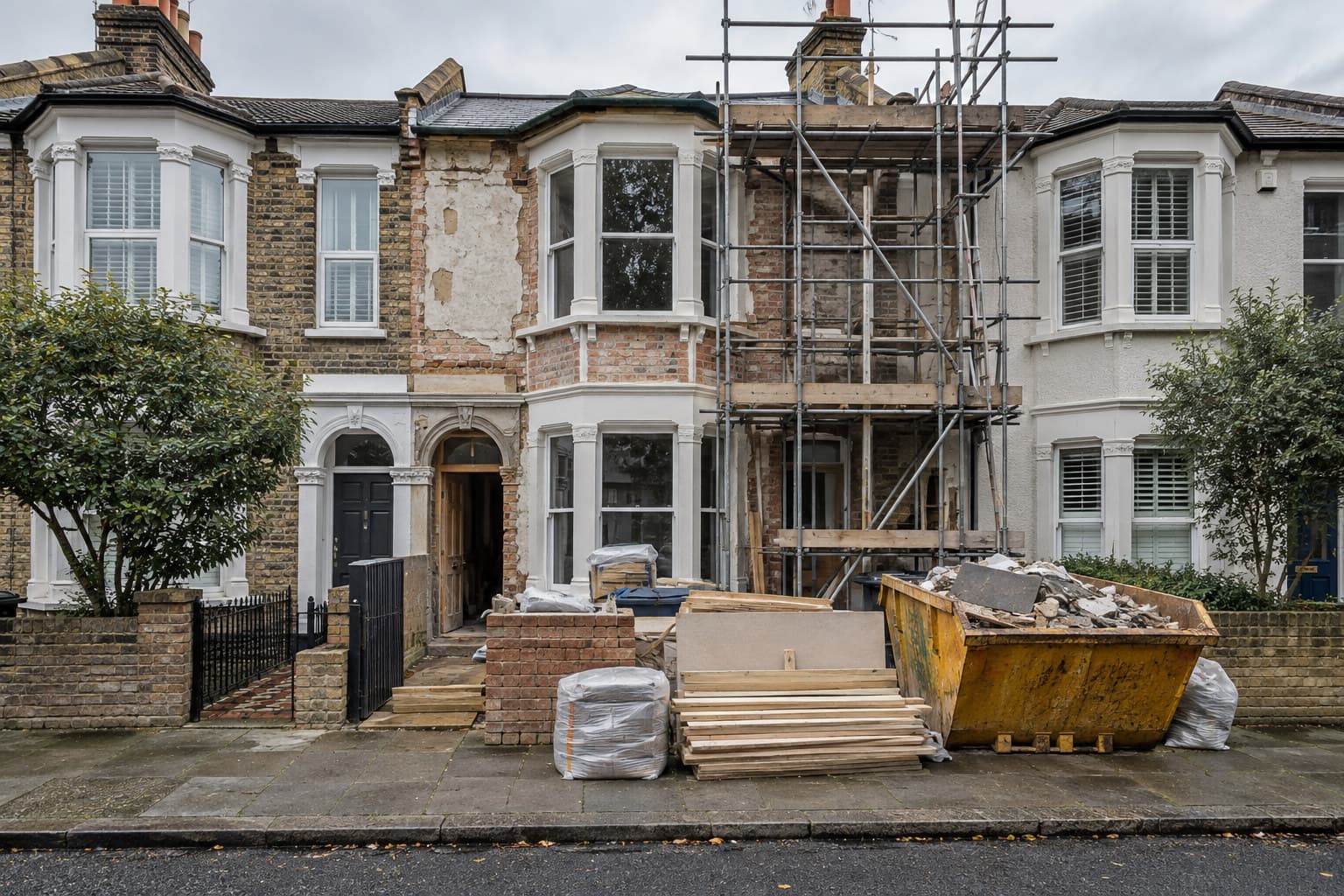 A UK house under renovation with scaffolding, building materials and a skip outside on an overcast day