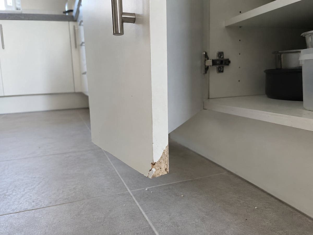 The lower outer corner of the white base-unit door is broken away, exposing the chipboard core.