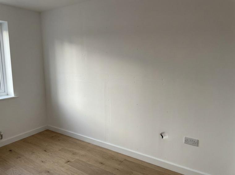 Plasterboard joints and screw dimples telegraphing through fresh white paint on a new build wall in raking daylight
